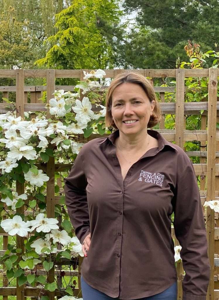 Image of the female company director of our family business in earls colne in front of our wooden trellis panels covered in white flowers. Jane is wearing her tarmec and croft fencing and gates brown uniform